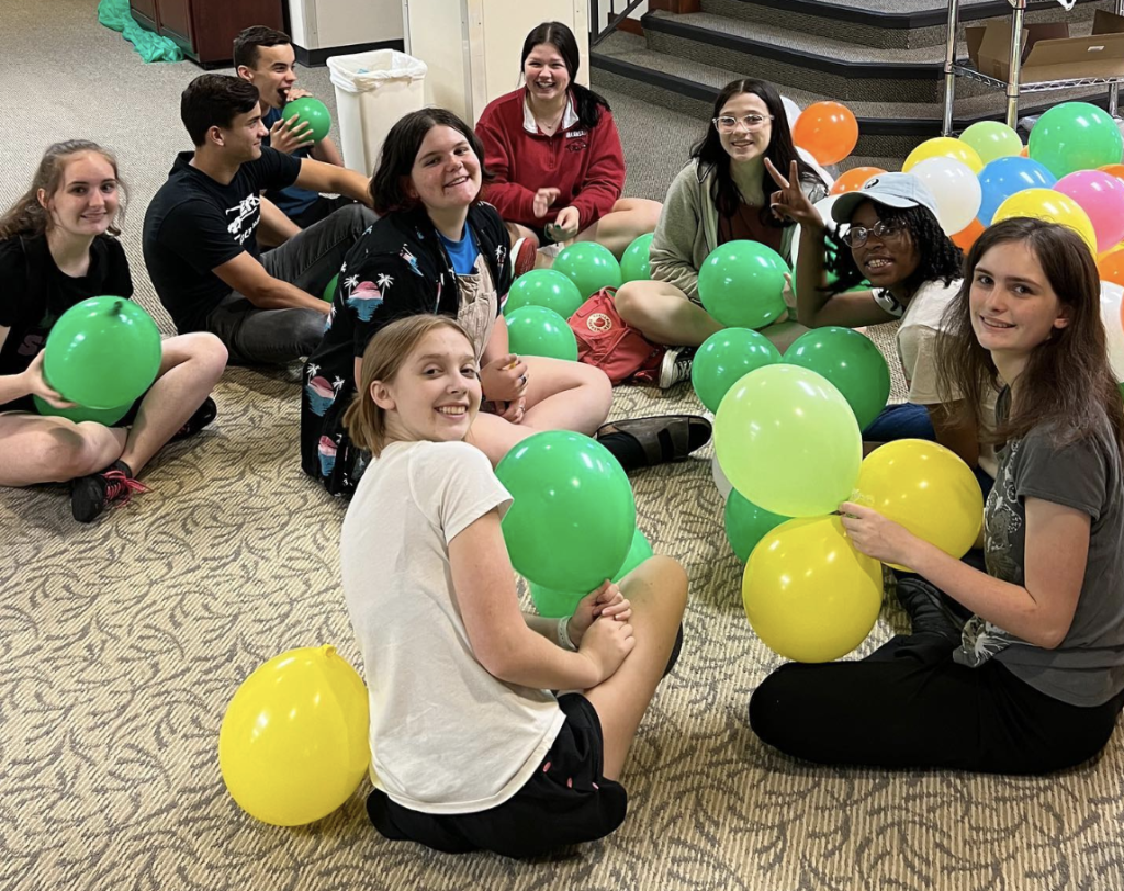 Photo of teens sitting on the floor holding yellow and green balloons.